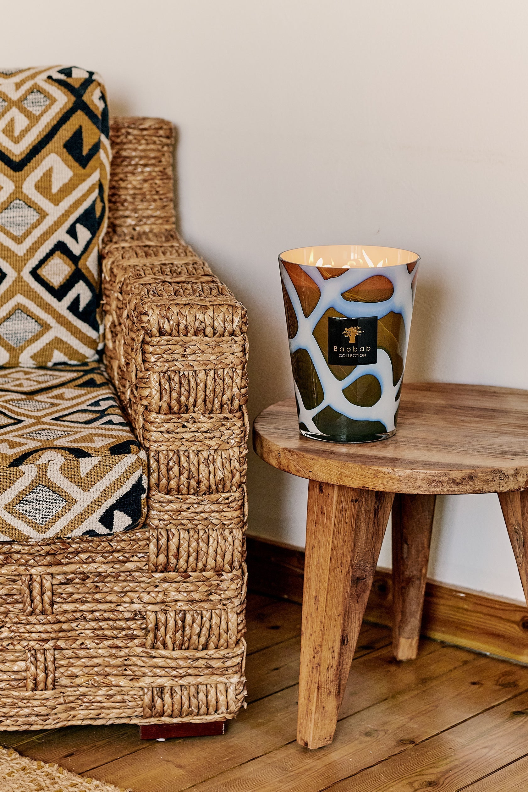 Scented Baobab Collection candle on a rustic wooden table next to a stylish woven chair and patterned cushion.