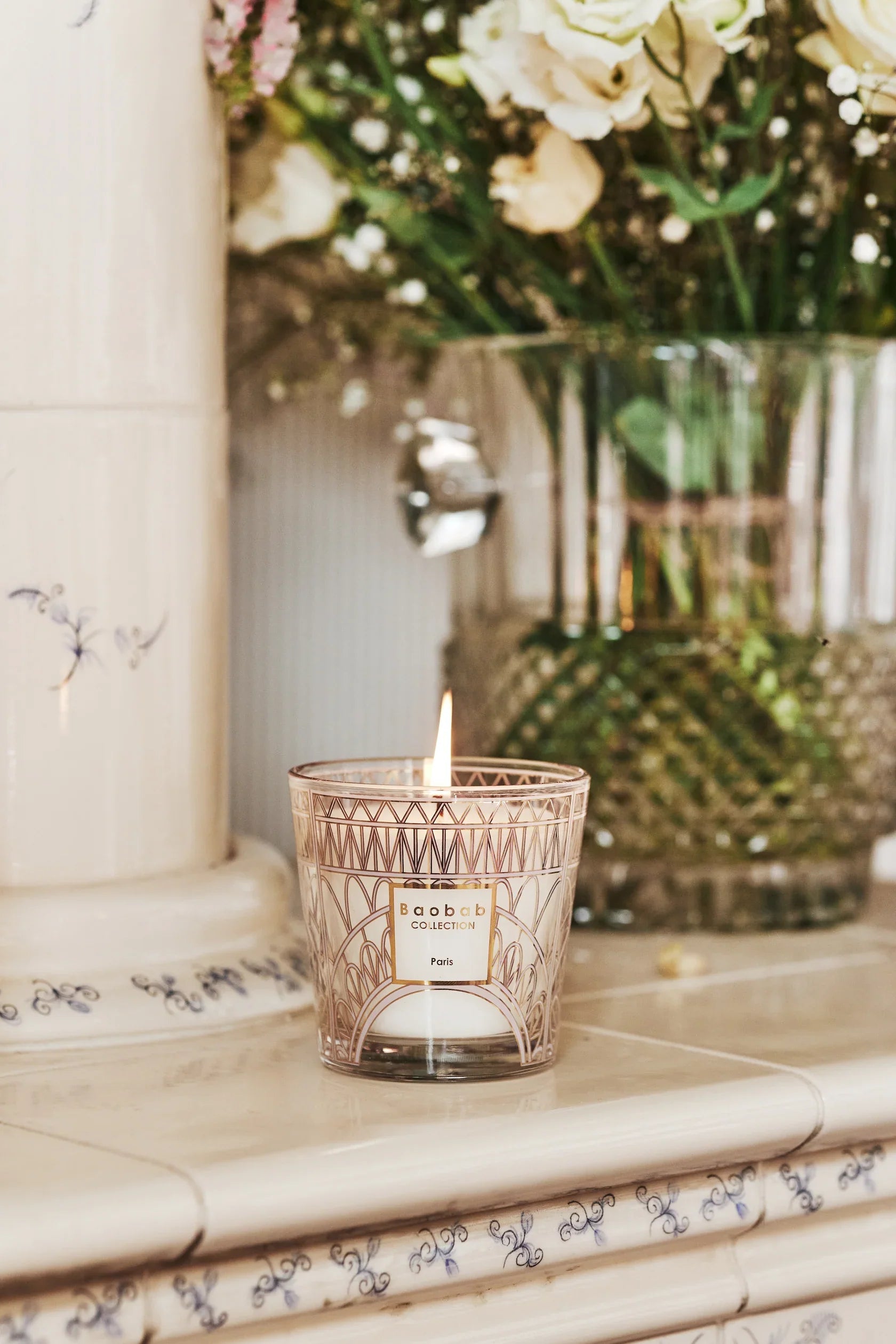 Baobab Collection scented candle with Eiffel Tower design, lit, placed on a mantel with flowers in the background.
