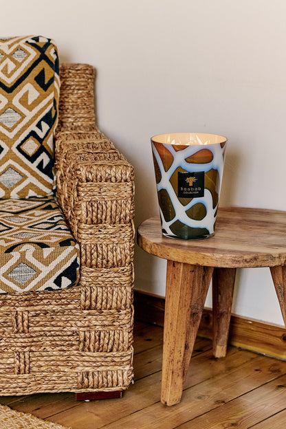 Scented Baobab Collection candle on a rustic wooden table next to a stylish woven chair and patterned cushion.