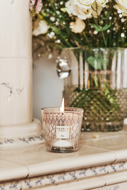 Baobab Collection scented candle with Eiffel Tower design, lit, placed on a mantel with flowers in the background.